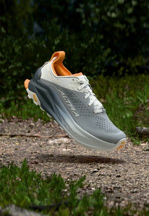 Šedobílá běžecká obuv Altra s oranžovým lemováním, zavěšená nad štěrkovou cestou obklopenou zeleným listovím.
