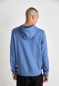 Hoodie in cotone blu con una texture morbida, dotato di cappuccio con cordino e maniche lunghe, mostrato da dietro su uno sfondo chiaro.
