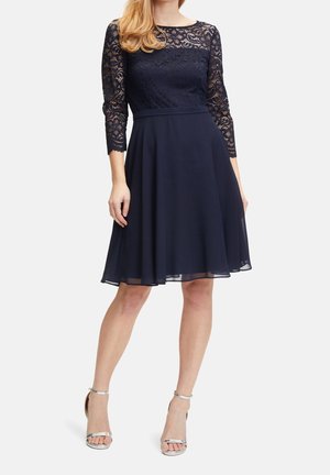 Kvinde iført en mørkeblå knælængde kjole med blonder og trekvartærmer samt sølvfarvede åbne ankelremssandaler, stående mod en hvid baggrund.