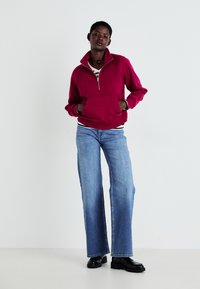 Sweatshirt zippé bordeaux avec un col haut, poche avant, associé à un jean noir à jambes larges et des chaussures à plateforme noires. Fond uni.