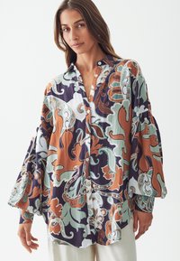 Camisa de botones con mangas largas y anchas en un patrón de paisley multicolor de verde, naranja y azul marino sobre un fondo claro. Tejido texturizado.