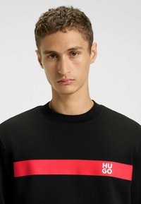 Felpa nera con una spessa striscia rossa orizzontale e il logo bianco "HUGO" sul lato sinistro del petto.