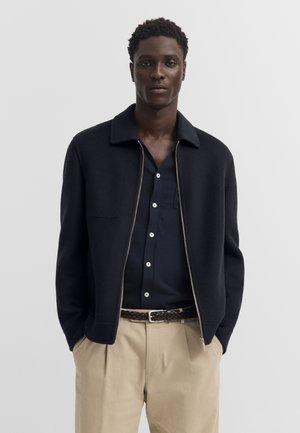 Mann, der eine marineblaue Jacke mit Reißverschluss über einem dunkelblauen, geknöpften Hemd, beige Hose und einen geflochtenen dunkelbraunen Gürtel trägt, steht mit den Händen in den Taschen.