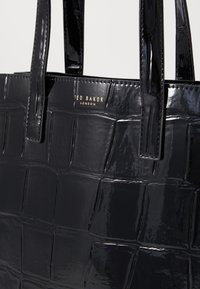Zwarte gepatenteerde faux-lederen handtas met een gequiltte textuur. Voorzien van twee korte handvatten en een gouden "Ted Baker London" logo.