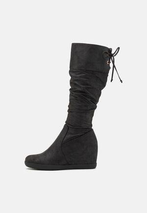 Czarna botka na koturnie do połowy łydki z luźną cholewką i sznurowaniem u góry na białym tle.