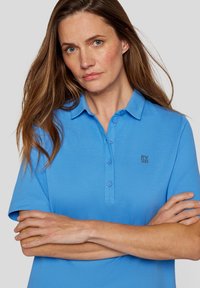 Helles blaues Polohemd mit Kragen, kurzen Ärmeln und durchgehender Knopfleiste. Mit kleinem Logo im linken Brustbereich.