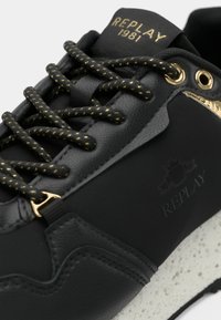 Gros plan sur une sneaker noire avec des accents dorés, des lacets noirs et dorés à motifs, une semelle texturée, et un logo "REPLAY 1981" sur la languette.