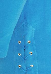 Blå bomullsskjorta med detaljer som en snörningsdesign med gyllene öglor och slät textur. Sid sömmarna framhäver geometrisk sömnad.