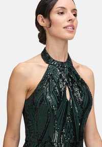 Frau in einem dunkelgrünen Paillettenkleid mit Halterneck, Schlüssel-Ausschnitt und geometrischem Muster, die Haare nach hinten gebunden vor einem schlichten Hintergrund.