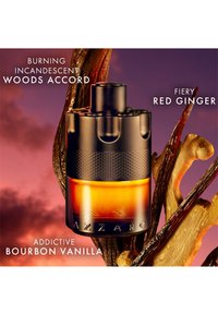 Azzaro-fles met gloeiend amberkleurige vloeistof, omringd door vanillepeulen, gemberwortel en rokerige houten elementen op een paars-oranje achtergrond.