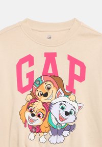 Μπεζ παιδικό T-shirt με μεγάλο ροζ κείμενο «GAP» και τρία χαμογελαστά κινούμενα κουτάβια που φορούν πολύχρωμο εξοπλισμό.