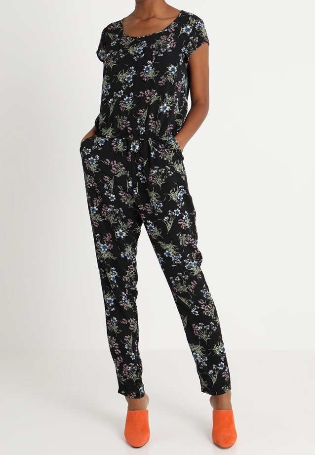 Femme portant une combinaison noire à fleurs avec des manches courtes et des poches, associée à des mules orange vif, debout devant un fond uni.