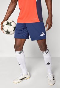 Άτομο που φορά μπλε σορτς, άσπρες ποδόσφαιρες κάλτσες και λευκά ποδοσφαιρικά παπούτσια κρατώντας μια λευκή και μαύρη ποδοσφαιρική μπάλα με αστέρια.