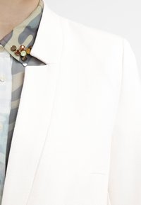 White blazer with a notched collar, paired with a patterned light blue and green camo shirt. The collar features decorative accents in gold and red.
