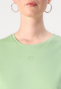 Šviesiai žalia apvalaus kaklo marškinėliai, pagaminti iš švelnios medžiagos, su tekstūruotu logotipu centre, papildyti didelėmis sidabrinėmis auskarų kilpomis.