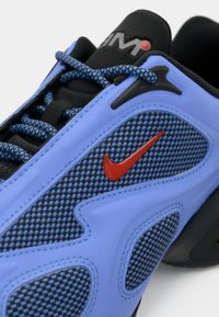 Zils un melns sporta apavu ar teksturētu acs virsu, izteiksmīgu sarkanu Nike logo, melnām šņorēm un melnu gumijas zolīti.