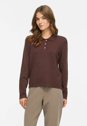 Femme aux cheveux bruns ondulés portant une chemise marron à manches longues avec col et boutons, et un pantalon beige, debout devant un fond clair uni.