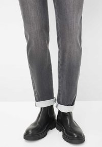 Gambe che indossano jeans grigi arrotolati e stivali Chelsea chunky neri in pelle su uno sfondo bianco.