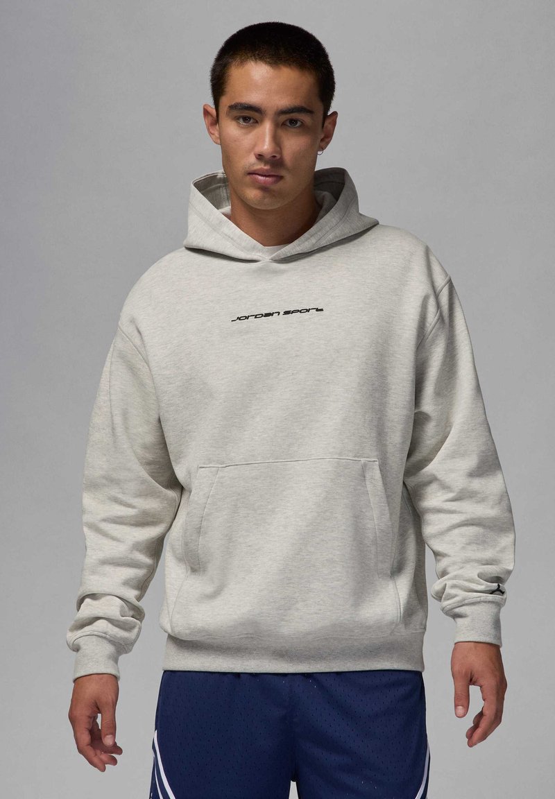 Young man wearing a light gray Jordan Sport hoodie and dark blue athletic shorts, standing against a plain gray background.