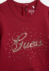 Abito borgogna con scollatura tonda, cerniera posteriore e logo "Guess" in paillettes argento, con paillettes sparse sulla parte anteriore.