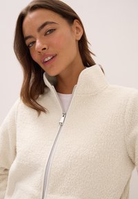 Jeune femme au teint clair et aux cheveux bruns portant une veste blanche à texture avec un col montant, souriant doucement.
