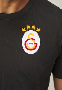 Maglia sportiva nera con un motivo testurizzato, un logo circolare bianco con accenti rossi e gialli, e cinque stelle gialle sopra il logo.