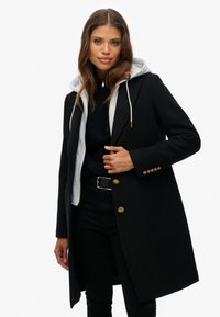 Zwarte wol-blend jas met een grijze gevoerde capuchon, gouden knopen en een getailleerd silhouet, gelaagd over een gestructureerde zwarte trui en broek.