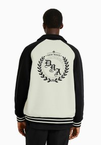 Svart och krämfärgad jacka med en logotyp som har "DHA" i kursiv, omgiven av lagerblad, randiga ärmslut och krage.