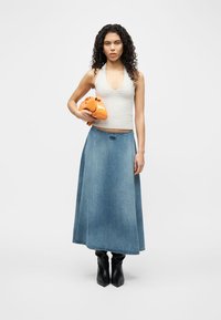 Γυναίκα με σγουρά μαλλιά φοράει γκρι αμάνικο τοπ και μακριά τζιν φούστα, κρατώντας μια πορτοκαλί τσάντα, στέκεται μπροστά σε μονόχρωμο λευκό φόντο.