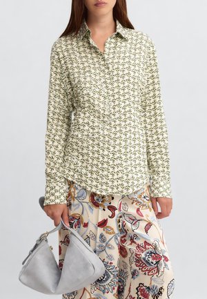 Donna che indossa una camicia bianca a maniche lunghe con motivi a foglia verde, pantaloni ampi floreali e tiene in mano una borsa in camoscio grigio chiaro.