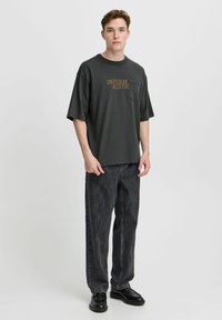 Tamno sivi oversized T-shirt s prednjim džepom i zlatnim tekstom. Uparen s crnim trapericama ravnog kroja i crnim cipelama. Glatka tekstura tkanine.