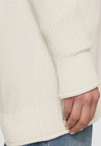 Gestrickter weißer Pullover mit strukturiertem Muster, gerippten Bündchen und Saum. Sichtbare Hand, die den unteren Rand greift und die Dicke des Stoffes betont.