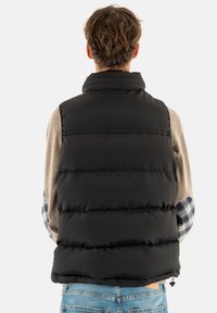 Chaleco negro acolchado con cuello alto, diseño acolchado, textura de canalé y dobladillo ajustable. Llevado sobre una camisa de manga larga beige y de cuadros.