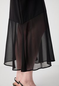 Vestito nero con un design a strati; la parte superiore è in tessuto consistente, mentre la parte inferiore è trasparente. Presenta un orlo che sfiora delicatamente la caviglia.