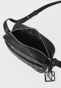 Schwarze Crossbody-Tasche mit einer strukturierten Lederoberfläche, Reißverschluss, Innenfutter mit einem Logomuster und einem dekorativen Metallschilder.