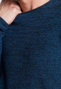 Pull bleu en tricot avec un motif texturé. Il présente un large décolleté et des manches longues. Le tissu semble doux et chaud.