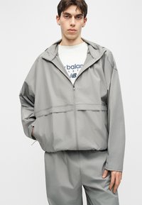 Jonge man in een grijze capuchonvest met rits en bijpassende broek, een wit New Balance-shirt dragend, staand met één hand in de zak.