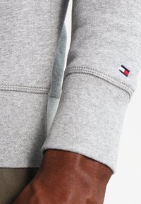 Detailní záběr na tmavou ruku v lehce šedé mikině s krátkým rukávem, na zápěstí se nachází malé logo v červené, bílé a námořnické modři.