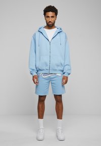 Light blue zip-up hoodie with front pockets, paired with matching shorts featuring a white script logo. Material is cotton blend. White sneakers.
