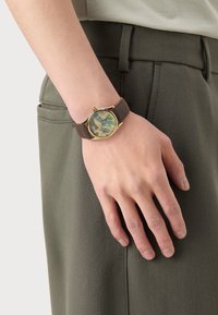 Montre avec bracelet en cuir brun et boîtier rond en acier doré, arborant un cadran à motifs bleu clair et beigne. Portée sur une main contre un pantalon vert olive.