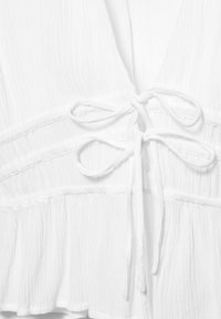 Tissu blanc texturé avec deux nœuds noués au centre, présentant des coutures froncées et un détail à volants au bas.
