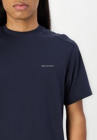 Tamsiai mėlynas apvalaus kaklo marškinėliai, pagaminti iš minkštos medžiagos, su trumpomis rankovėmis ir mažuoju "BELSTAFF" logotipu kairėje krūtinės pusėje.