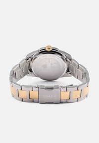 Reloj de acero inoxidable y tono dorado con una correa de malla plateada, corona dorada y parte trasera de la caja grabada. Resistente al agua hasta 50 metros.