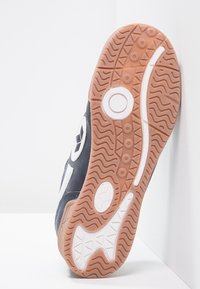 Baskets de sport noires avec une semelle en caoutchouc texturée. Accents blancs et logo sur le côté. Le profil montre des rainures et des motifs distincts sur la semelle extérieure.