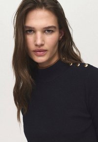 Navy knit sweater with a high neckline, featuring three gold-button accents on the left shoulder. Smooth texture and fitted design.