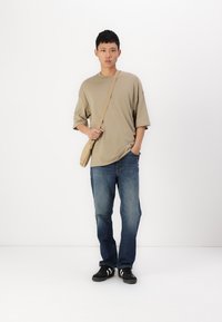 Béžové oversized tričko, modré džínové kalhoty a černé boty s bílými pruhy; model drží světle hnědou tašku přes rameno a stojí na bílém pozadí.