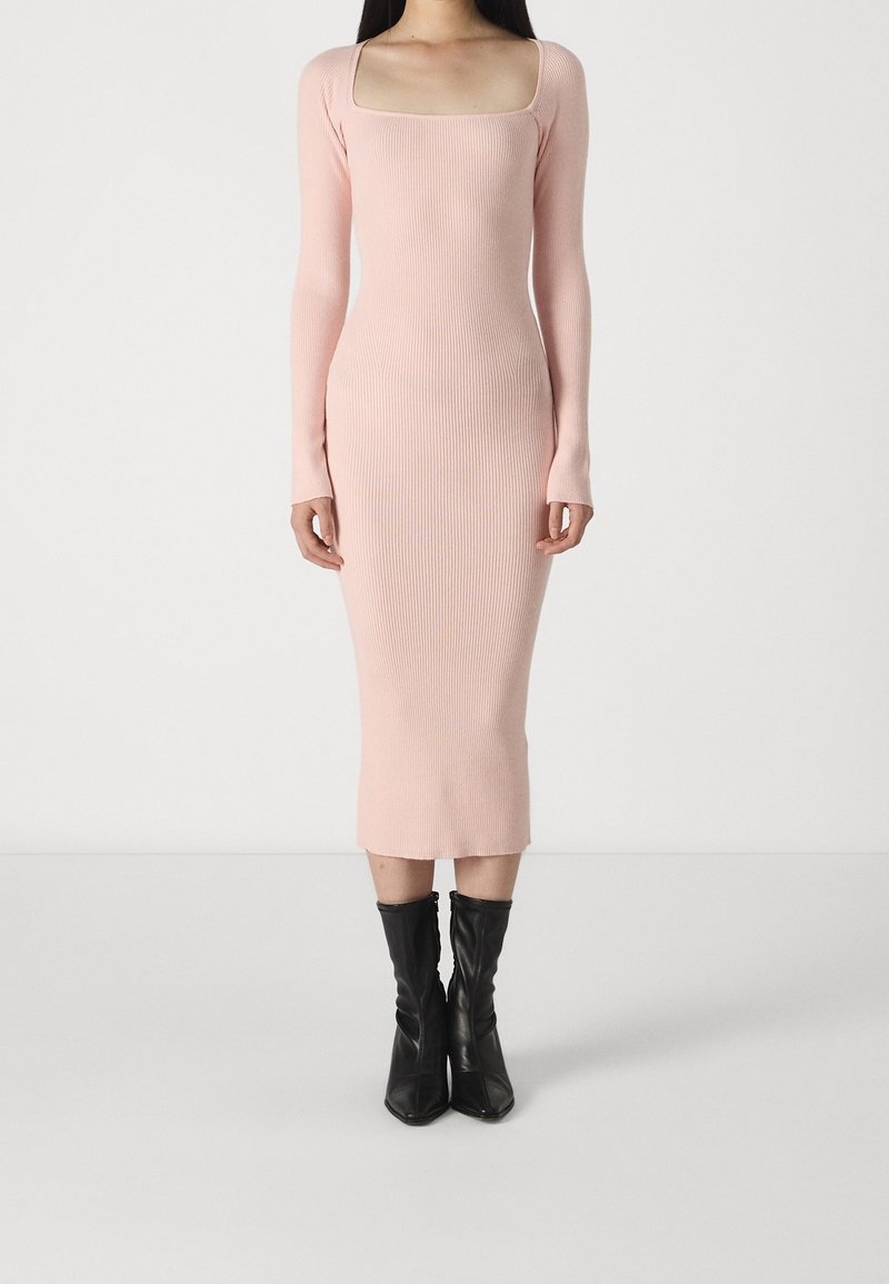 Femme portant une robe midi côtelée rose pâle à manches longues avec un décolleté carré, associée à des bottines noires à bout pointu.