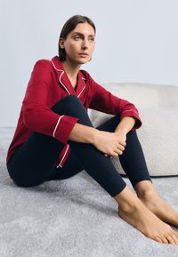 Červená pyžamová blúzka s bielym lemem, spárovaná s čiernymi legínami. Modelka sedí na svetlošedej textúrovanej koberci.