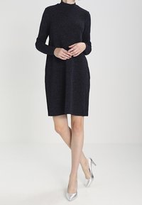 Schwarzes langärmeliges Kleid mit hohem Ausschnitt, aus strukturiertem schimmerndem Stoff. Knielang mit lockerer Passform; kombiniert mit silbernen High Heels.