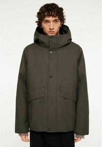 Mörkgrön, huva jacka med lös passform, som har tryckknappar, två framfickor och ribbade manschetter synliga under ärmarna på jackan.